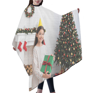 Personality  Happy Girl In Party Cap Holding Gift Box Near Blurred Christmas Tree And Fireplace At Home  Hair Cutting Cape