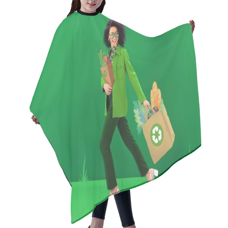 Personality  smiling and stylish african american woman walking with illustrated shopping bags with recycling signs on green background  hair cutting cape