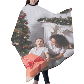 Personality  Girl With Open Mouth Holding Big Gift Box Near Mother And Festive Pine At Home Hair Cutting Cape