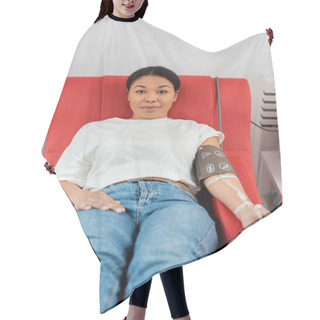 Personality  Positive Multiracial Woman With Pressure Cuff, Transfusion Set And Medical Rubber Ball Sitting On Medical Chair And Looking At Camera During Blood Donation In Clinic Hair Cutting Cape