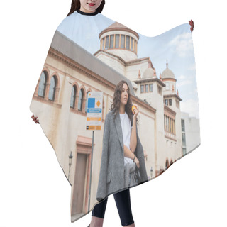 Personality  Young Curly Woman In Grey Casual Jacket Holding Fresh Orange And Leash While Looking Away With Blurred Historical Landmark At Background In Barcelona, Spain, Ancient Building  Hair Cutting Cape