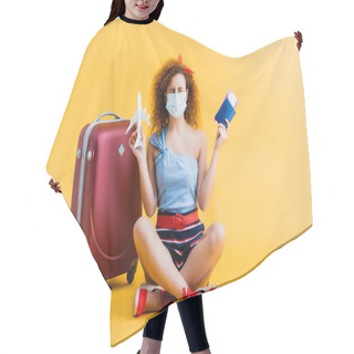 Personality  Curly Young Woman In Medical Mask Holding Passport And Toy Plane While Sitting Near Suitcase On Yellow  Hair Cutting Cape