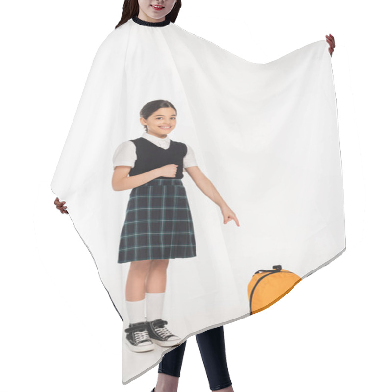 Personality  cheerful girl in school uniform standing and pointing at backpack on white background, full length hair cutting cape