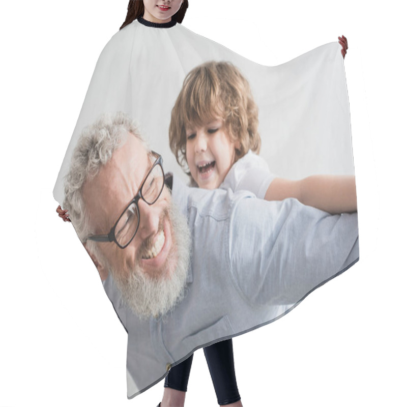 Personality  Cheerful granddad playing with blurred grandson at home hair cutting cape