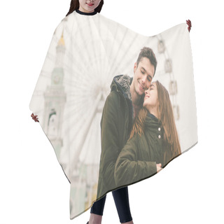 Personality  Theme Love And Holiday Valentines Day. Pair Of Caucasian Heterosexual Lovers In Winter Together Gloomy Weather Embrace Against Background Of Ferris Wheel In Town Square. The Guy Gently Hugs The Girl. Hair Cutting Cape