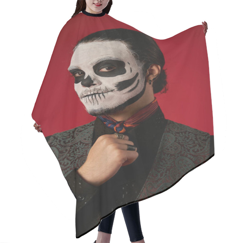 Personality  portrait of man in catrina makeup and stylish attire looking at camera on red, ria de los muertos hair cutting cape
