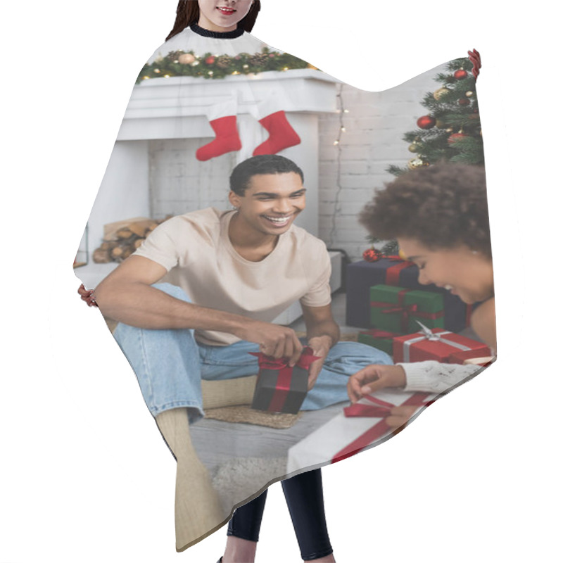 Personality  smiling african american man looking at girlfriend opening gift box near christmas tree hair cutting cape