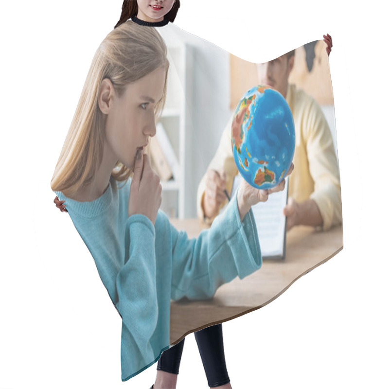 Personality  selective focus of thoughtful young woman looking at globe while sitting near travel agent hair cutting cape