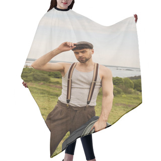 Personality  Trendy Bearded Man In Suspenders Touching Newsboy Cap And Holding Jacket While Looking At Camera With Rural Landscape At Background, Fashion-forward In Countryside Hair Cutting Cape