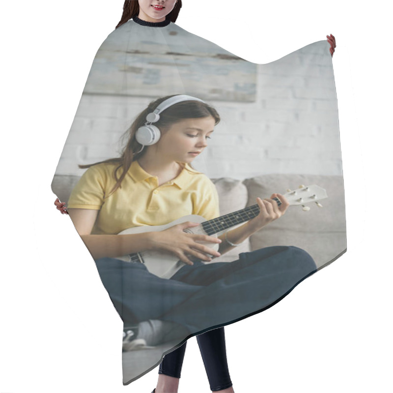 Personality  Preteen Girl In Wired Headphones Playing Ukulele While Sitting With Crossed Legs On Couch  Hair Cutting Cape