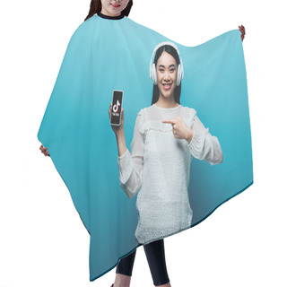 Personality  KYIV, UKRAINE - JULY 15, 2019: Smiling Asian Woman With Headphones Pointing With Finger At Smartphone With TikTok Icon On Blue Background Hair Cutting Cape