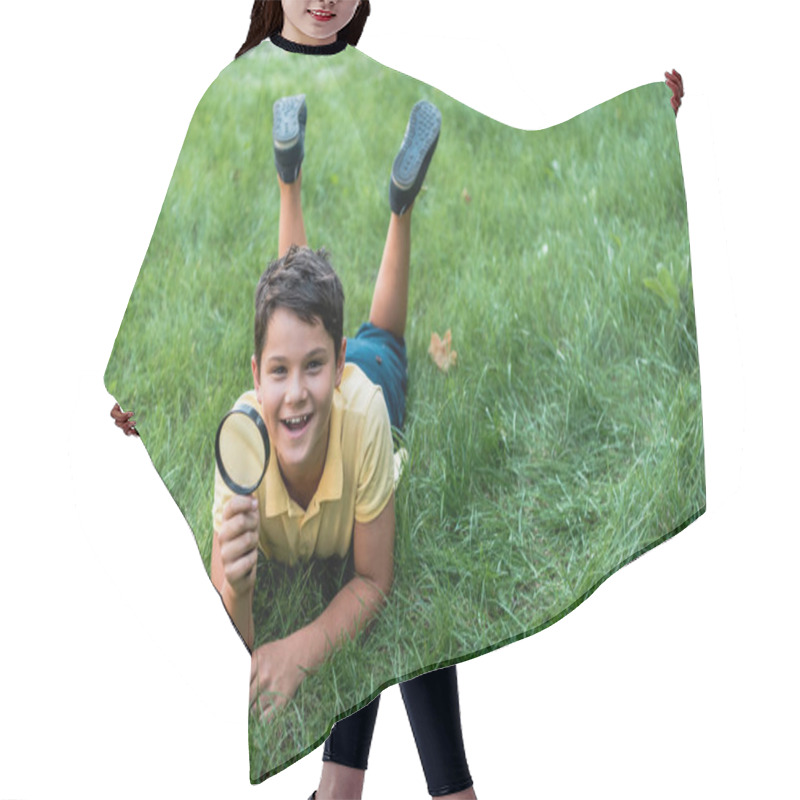 Personality  cheerful boy lying on grass and holding magnifier  hair cutting cape