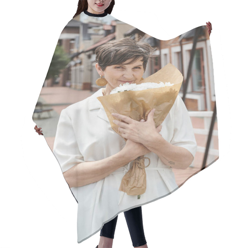 Personality  elderly woman holding bouquet of flowers and looking at camera, urban backdrop, summer, white outfit hair cutting cape