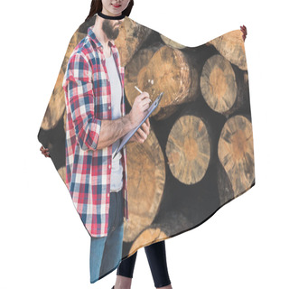 Personality  Cropped Shot Of Bearded Lumberjack In Checkered Shirt Writing In Clipboard On Logs Background  Hair Cutting Cape