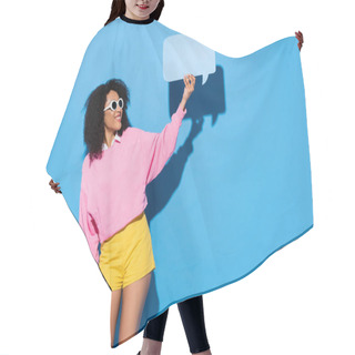 Personality  Pleased African American Woman In Pink Sweatshirt And Yellow Shorts Showing Empty Speech Bubble On Blue Hair Cutting Cape