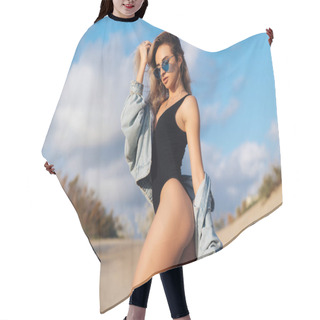 Personality  Sexy Girl On The Beach In A Black Swimsuit And Blue Sunglasses With A Denim Jacket In Her Hands. Beautiful Model With A Sports Figure Walks Near The Ocean On Vacation, Enjoying Nature. Hair Cutting Cape