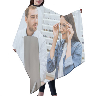 Personality  Happy Woman Choosing Eyeglasses And Looking At Mirror While Male Oculist Standing Near With Laptop In Optics  Hair Cutting Cape