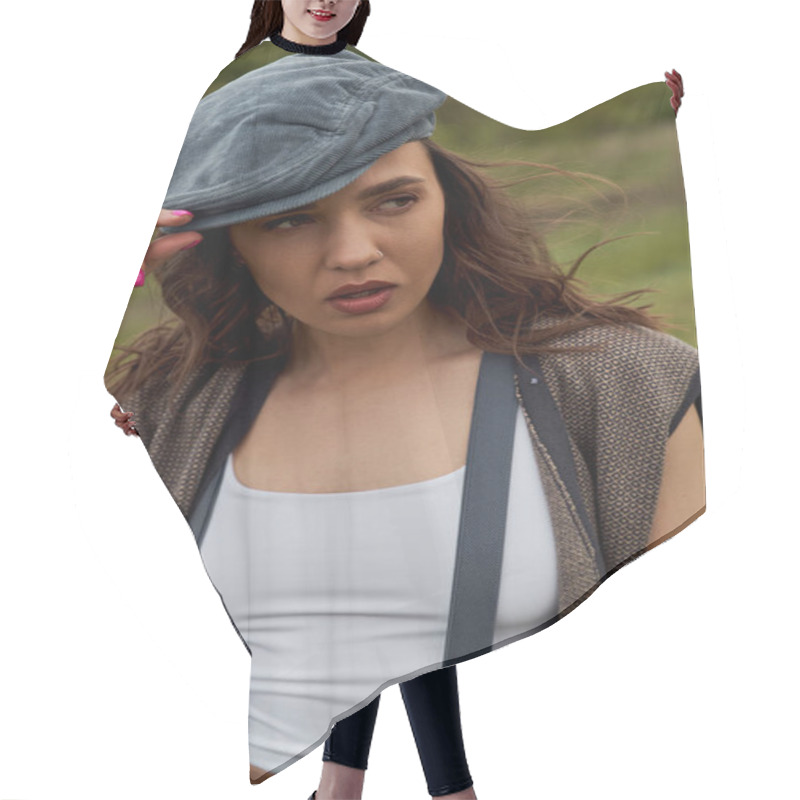 Personality  Portrait of fashionable brunette woman in vest and vintage suspenders touching newsboy cap and looking away while standing at blurred nature, fashion-forward in countryside hair cutting cape