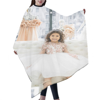 Personality  Cheerful Middle Eastern Girl With Curly Hair Sitting In Floral Dress On White Couch Inside Of Luxurious Wedding Salon, Smiling Kid, Tulle Skirt, Bridal, Blurred Mannequin On Background  Hair Cutting Cape