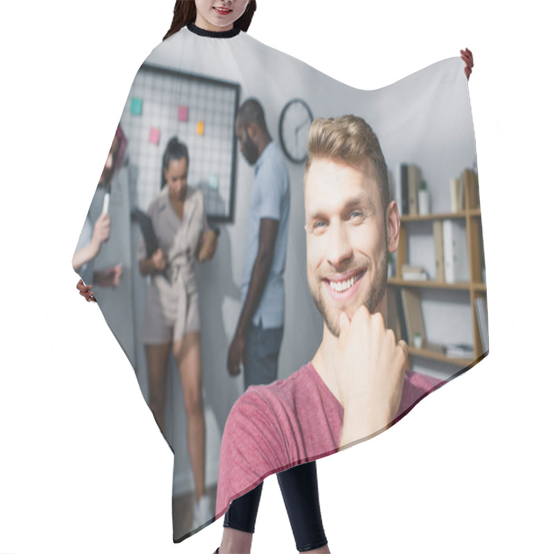 Personality  Businessman With Hand Near Chin Looking At Camera While Multicultural Coworkers Working In Office At Background  Hair Cutting Cape