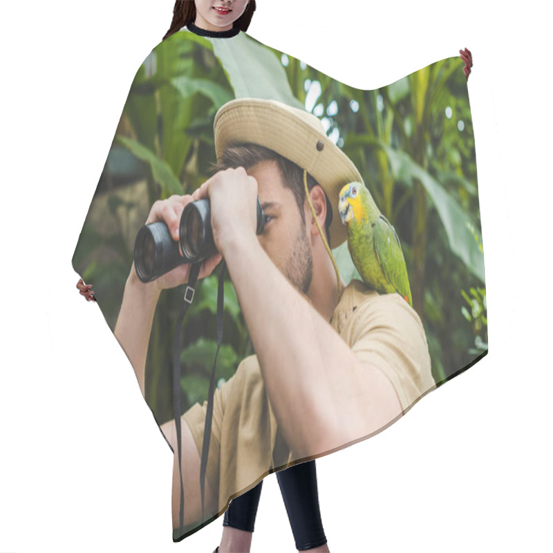 Personality  attractive young man with parrot on shoulder looking through binoculars in jungle hair cutting cape