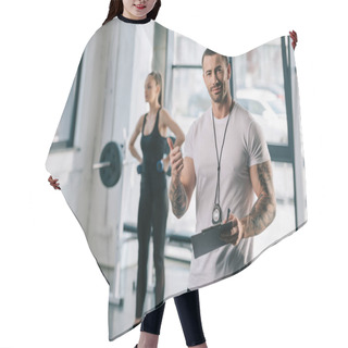 Personality  Personal Trainer With Clipboard Doing Thumb Up Gesture And Young Sportswoman Exercising With Dumbbells Behind At Gym Hair Cutting Cape