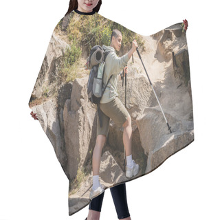Personality  Young Short Haired Female Traveler In Casual Clothes With Backpack Holding Trekking Poles While Standing Near Hill With Stones At Background During Summer, Translation Of Tattoo: Love Hair Cutting Cape