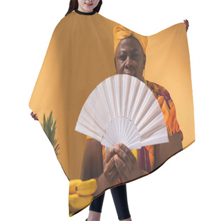 Personality  Serious Middle Aged African American Woman Sitting Near Fruits And Holding Fan On Orange Hair Cutting Cape
