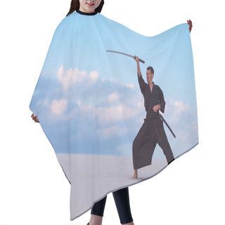 Personality  Focused Man, In Traditional Japanese Clothes, Is Practicing Martial Arts In The Desert With A Japanese Sword - A Katana During Sunset On The Background Of Blue Sky And Clouds. Hair Cutting Cape
