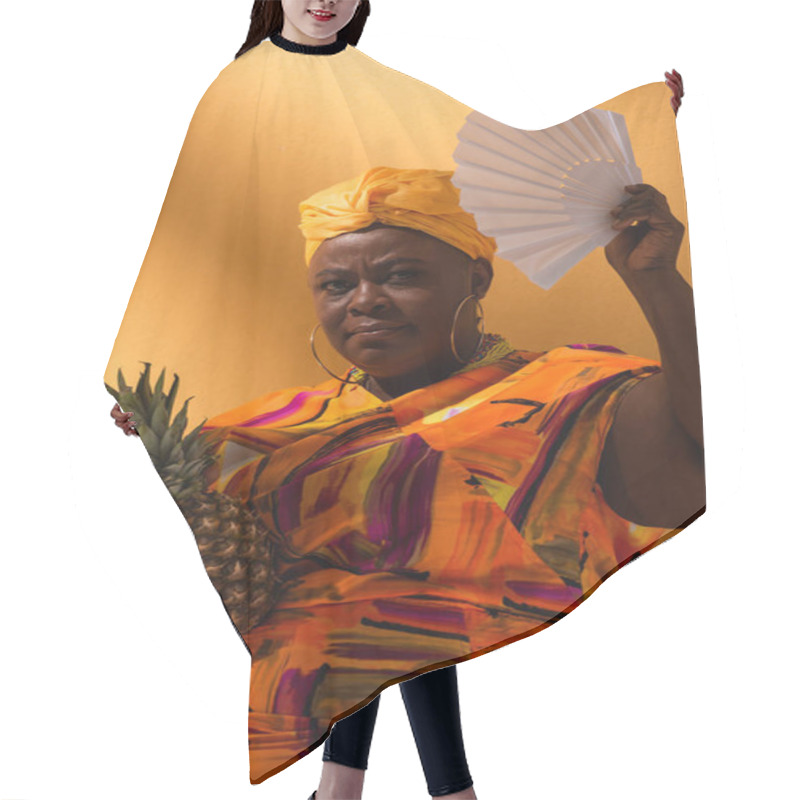 Personality  Serious Middle Aged African American Woman Holding Pineapple And Waving With Fan On Orange Hair Cutting Cape