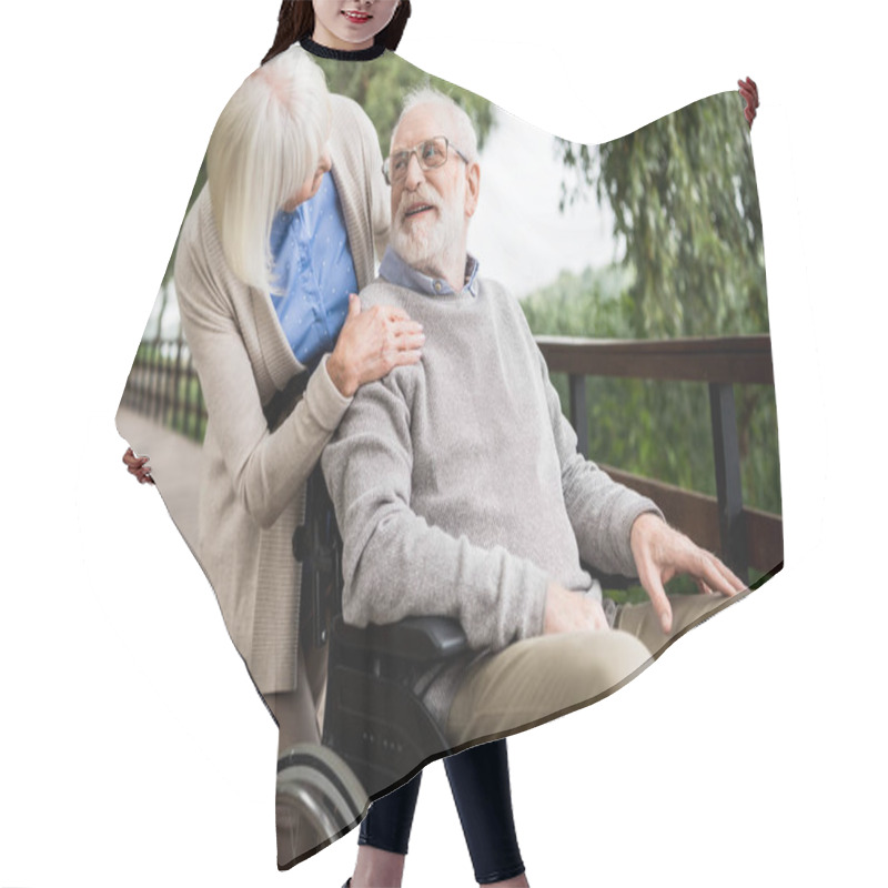Personality  senior woman looking at smiling husband in wheelchair while holding hand on his shoulder hair cutting cape