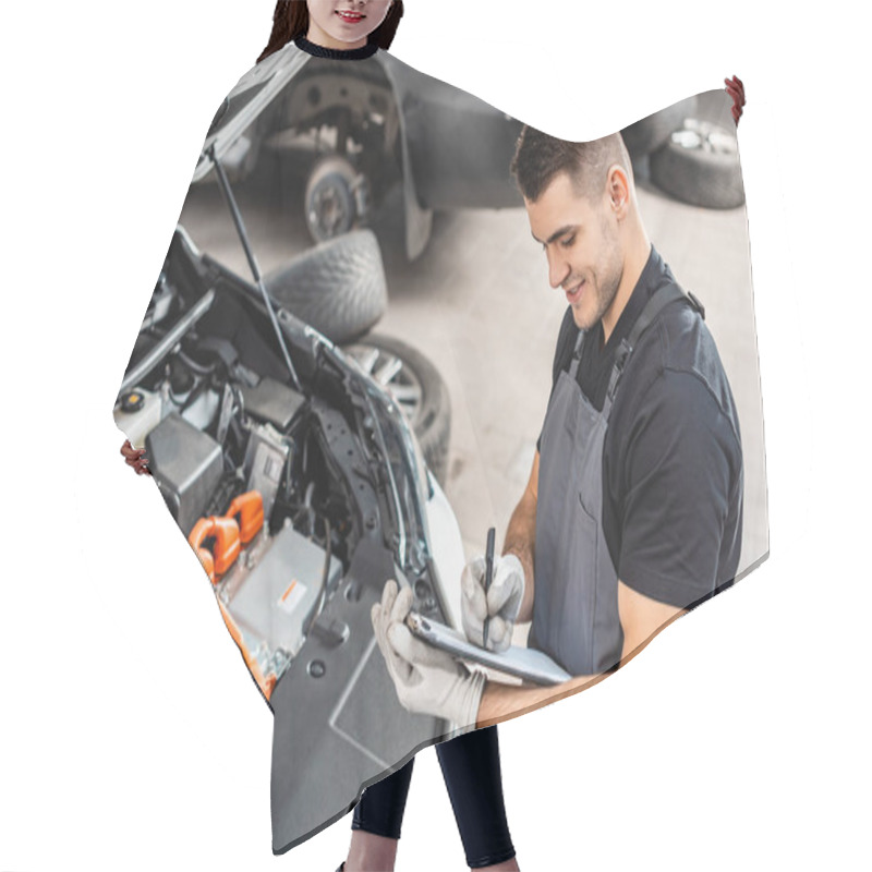 Personality  smiling mechanic writing on clipboard while inspecting car engine compartment hair cutting cape