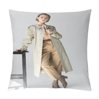 Personality  Full Length Of Young Woman In Glasses, Trench Coat And Scarf Leaning On Stool While Posing On Grey Pillow Covers