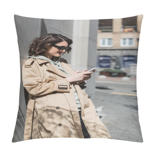 Personality  Cheerful And Trendy Woman With Wavy Brunette Hair And Tattoo, In Dark Sunglasses And Beige Trench Coat Standing Near Grey Building And Browsing Internet On Mobile Phone On Street In European City Pillow Covers