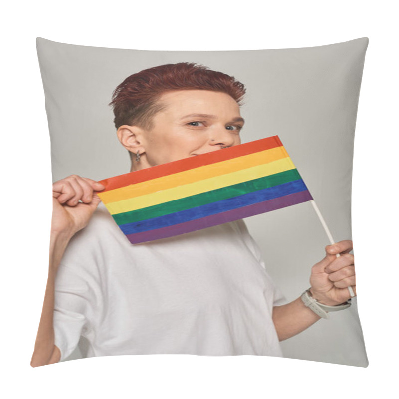 Personality  redhead queer model in white t-shirt posing with small LGBT flat near face looking at camera on grey pillow covers