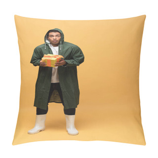 Personality  Confused Mixed Race Man In Raincoat And Rubber Boots Holding Gift Box Isolated On Yellow Pillow Covers