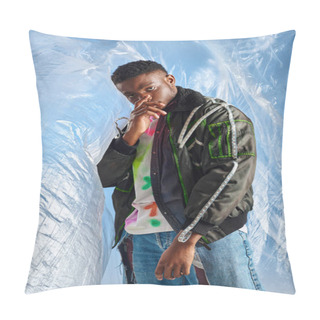Personality  Confident Young Afroamerican Model Looking At Camera While Covering Face And Posing In Outwear Jacket With Led Stripes And Ripped Jeans Glossy Cellophane On Blue Background, Sustainable Lifestyle  Pillow Covers