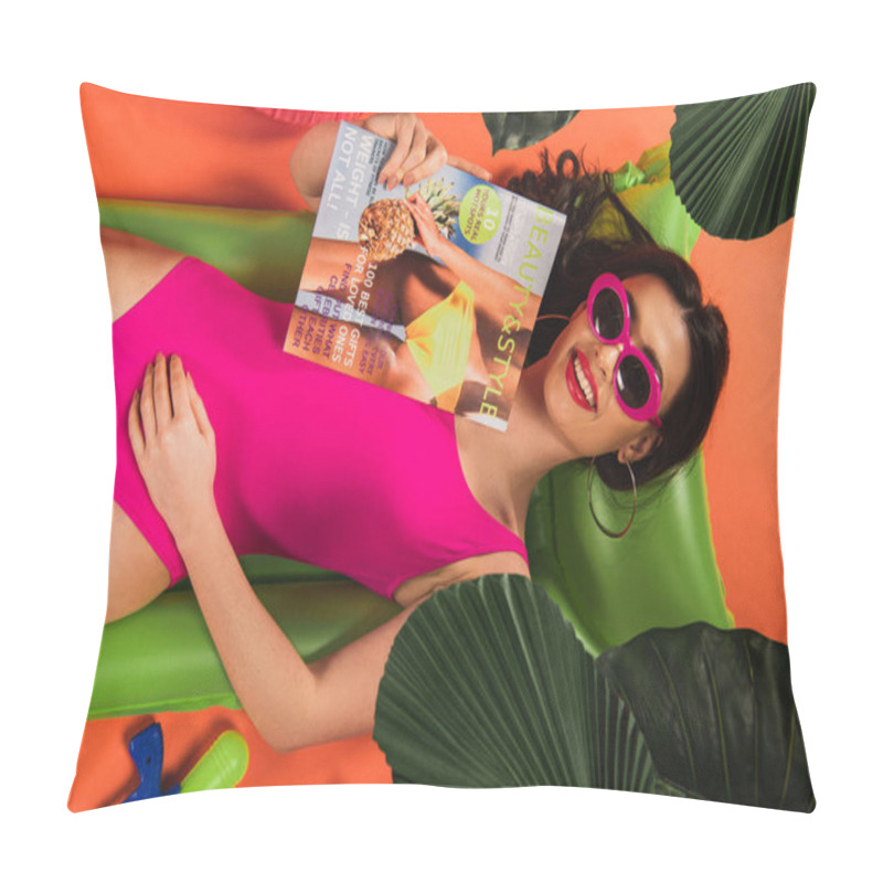 Personality  top view of happy young woman in sunglasses and swimsuit lying on inflatable mattress with magazine near green palm leaves on orange pillow covers