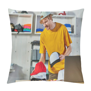 Personality  Cheerful Young Redhead Craftsman Holding Snapbacks While Working Near Cloth Samples And Blurred Laptop On Wooden Table In Print Studio, Small Business Resilience Concept Pillow Covers