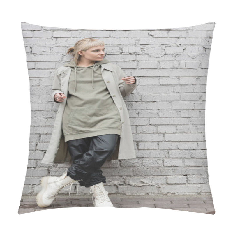 Personality  fashonable young woman with makeup, blonde hair, bangs, in stylish outfit, long hoodie, coat, black leather pants and beige boots standing near grey brick wall and looking away pillow covers
