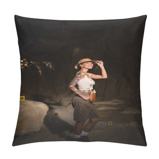Personality  Sexy Tattooed Archaeologist Adjusting Safari Hat And Smiling At Camera Near Numbered Cards In Dark Cave Pillow Covers