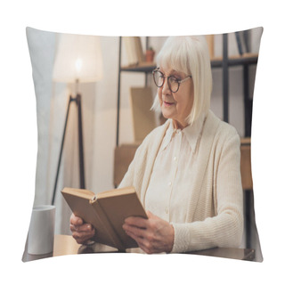 Personality  Thoughtful Senior Woman In Glasses Sitting At Table And Reading Book At Home Pillow Covers