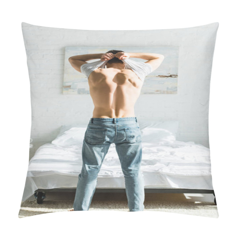 Personality  man in jeans standing backwards near bed and taking off white t-shirt in bedroom pillow covers