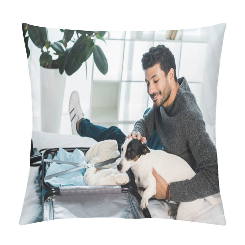 Personality  handsome and smiling bi-racial man stroking jack russell terrier and sitting near suitcase pillow covers