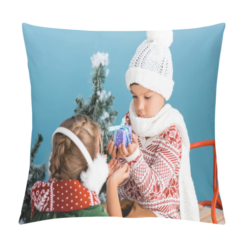 Personality  selective focus of boy in knitted hat holding present while sitting on sleight near girl in winter earmuffs on blue pillow covers
