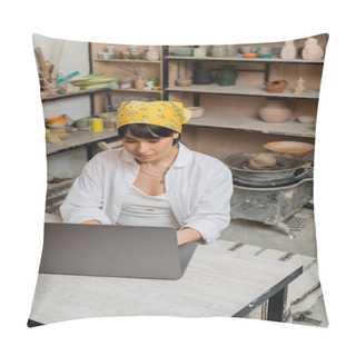Personality  High Angle View Of Young Asian Female Artisan In Headscarf And Workwear Using Laptop While Sitting At Table And Working In Blurred Ceramic Workshop, Pottery Artist Showcasing Craft Pillow Covers