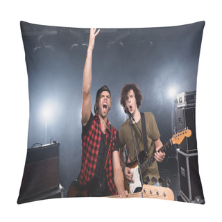 Personality  KYIV, UKRAINE - AUGUST 25, 2020: Rock Band Musicians Shouting While Holding Guitars Near Microphone Rack And Combo Amplifiers With Backlit On Background Pillow Covers