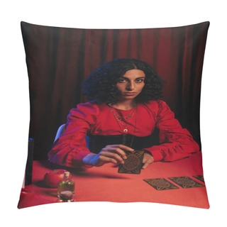 Personality  KYIV, UKRAINE - JUNE 29, 2022: Young Brunette Medium With Tarot Card Looking At Camera On Dark Background With Red Drape Pillow Covers