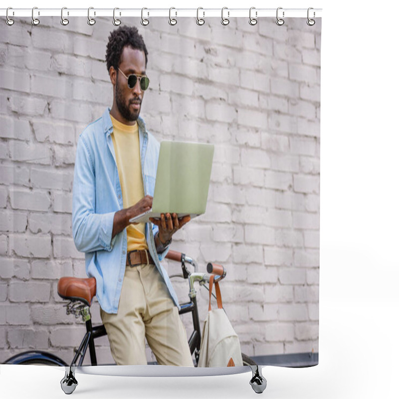 Personality  attentive, stylish african american man using laptop while standing by brick wall near bicycle shower curtains