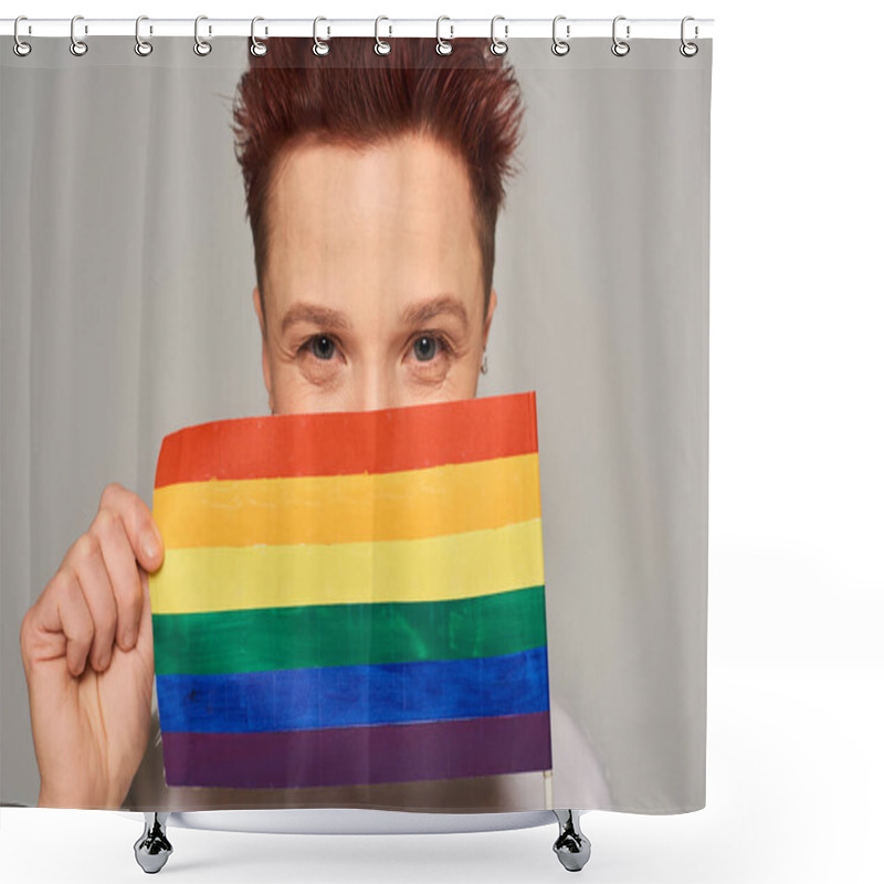 Personality  joyful redhead queer person obscuring face with small LGBT flag and looking at camera on grey shower curtains